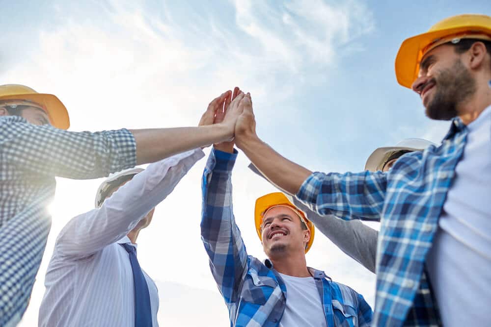 Depositphotos_82381250_S (1) Five Smartway Builders workers in hard hats in a circle, high fiving to celebrate a positive outcome.